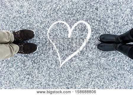 Men and women's boots standing at abstract heart symbol on asphalt covered gritty snow surface. Rough snowy. Cold Winter. Top view. Relations concept