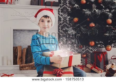 Christmas miracle. Cute happy boy in santa hat unwrap magic present on holiday morning in beautiful room. Male child open Xmas gift near decorated fir tree and fireplace. Winter holidays concept