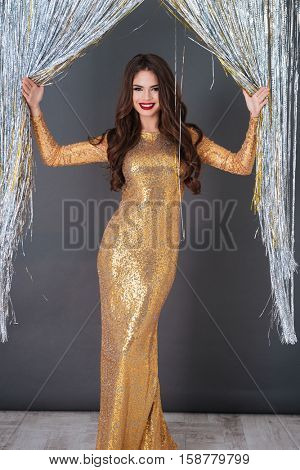 Cheerful gourgeous young woman in golden dress standing over sparkling decorations background