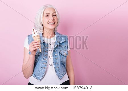 Good mood. Delighted pretty senior woman smiling and holding an icecream while standing against isolated pink background.