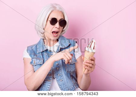 My favorite icecream. Expressive senior pretty woman opening her mouth and pointing on icecrem in her hand while standing against isolated pink background.