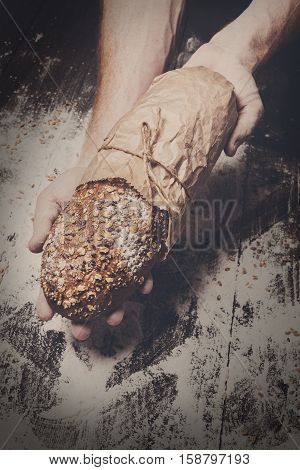 Warm fresh rye whole grain bread, wrapped in craft paper. Healthy baking and cooking concept background. Hands carefully hold loaf on wooden table background, sprinkled with flour. Soft toning