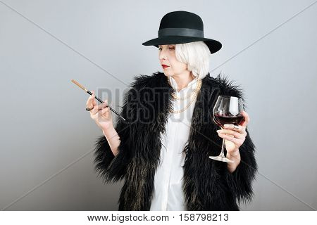 Aristocratic lifestyle. Presentable beautiful senior woman drinking wine and smoking cigarette while standing against isolated gray background.