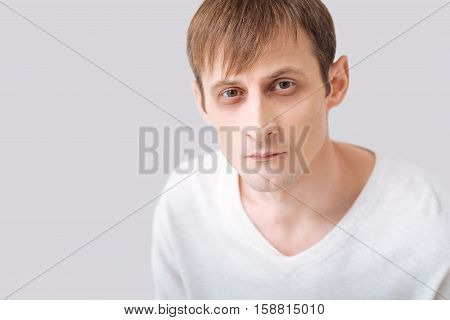 Fixed gaze. Serious thoughtful young man wearing a white pullover and looking at you while standing against the grey background