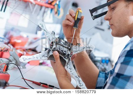 Service and technology. Handsome young delighted man repairing drone while using screvdriver and sitting in a workroom.