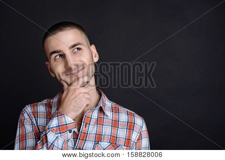 Positive thoughts. Young brunette feeling happiness staring aside touching his chin wearing checked shirt