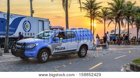 26 November 2016. Police car with policeman sitting in it on the background of a beautiful sunset with the orange sun, green palms and a big truck in Ipanema beach, Rio de Janeiro, Brazil