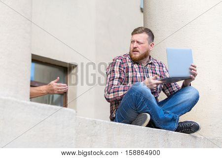Man sitting on the ledge of the building facade. It uses a laptop a wireless communication on the Internet. For a guy whose stretches a hand distracted from affairs
