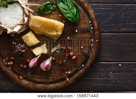 Cheese delikatessen background closeup on rustic wood. Wooden desk with parmesan, camembert and brie cuts decorated with garlic, pomegranate and basil, top view image
