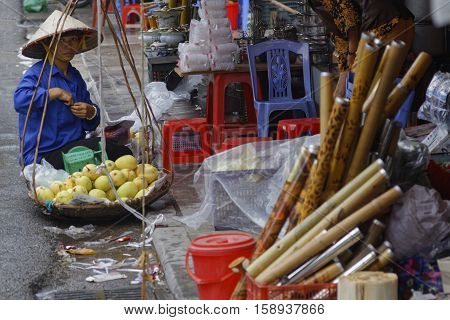 HANOI VIETNAM October 31 2016 : A woman transports fruits on balanced baskets in front of assorted ustensils of a general store straight from the street in Hanoi Vietnam.