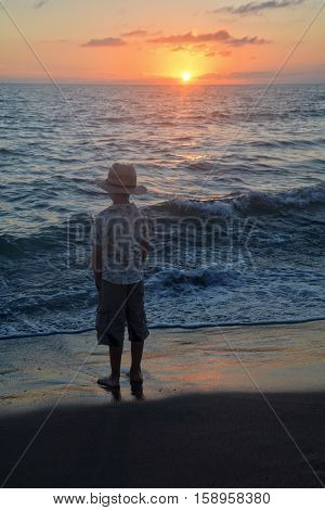 Small boy stands on the beach at the sundown, hands in the trouser pockets and looks to the horizon