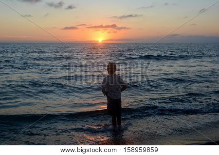 Small boy stands in the water on the beach at the sundown and looks as the sun