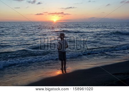 Small boy stands in the water on the beach at the sundown and looks as the sun