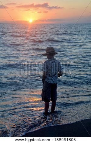 Small boy stands in the water on the beach at the sundown and looks in the water