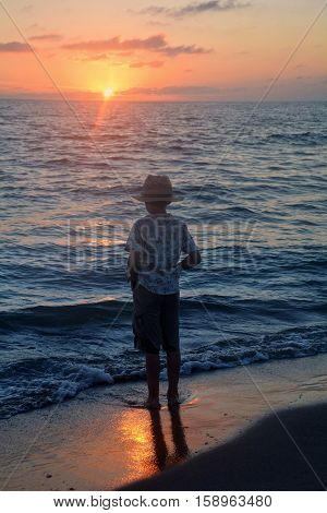 Small boy stands in the water on the beach at the sundown and looks as the sun