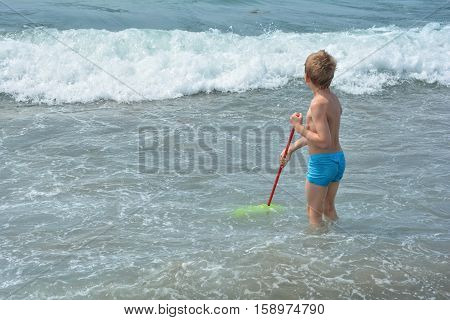 Small blond boy with blue swimming trunks stands with a fishing-net in the water and waits for the sea - wave