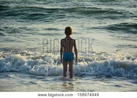 Small blond boy with blue swimming trunks stands in the water before a wave and looks to the sea