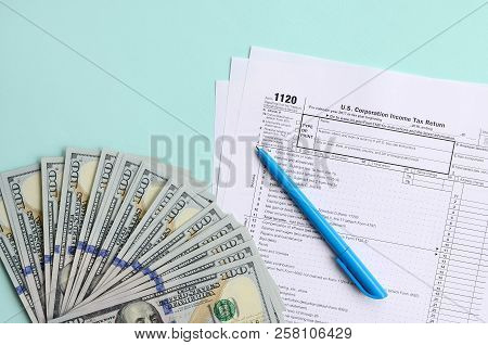 1120 Tax Form Lies Near Hundred Dollar Bills And Blue Pen On A Light Blue Background. Us Corporation