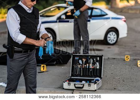 Partial View Of Male Police Officer In Sunglasses Putting On Latex Gloves While His Colleague Standi