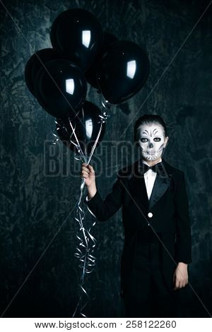 Portrait of a child boy with carnival make-up of a skeleton wearing elegant tail-coat. Studio shot over black background. Halloween. Dia de los muertos. The Day of the Dead.