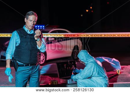 Mature Male Police Officer Talking On Radio Set While Criminologist Collecting Evidence At Crime Sce