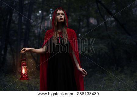 Elegant Mystic Girl In Red Cloak With Kerosene Lamp Walking In Dark Woods