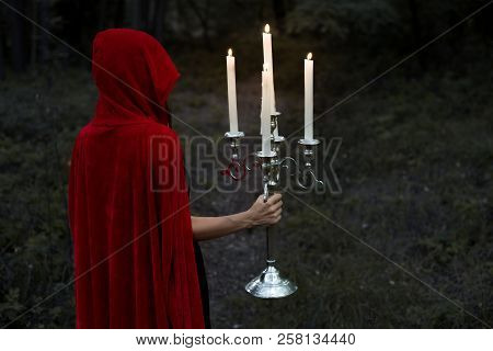 Mystic Girl In Red Cloak Holding Candelabrum With Flaming Candles In Dark Woods