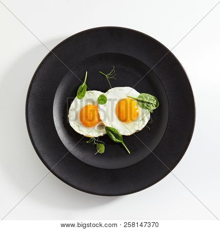 Elegant Restaurant Plate of Fried Eggs Decorated with Fresh Spinach Leaves and Young Pea Shoots on Flat Black Plate. Macro Photo of Healthy Nutritious Breakfast Isolated on White Background
