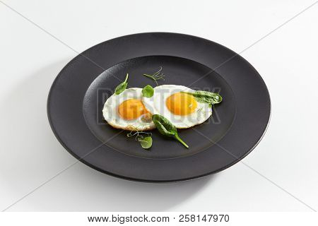Elegant Restaurant Plate of Fried Eggs Decorated with Fresh Spinach Leaves and Young Pea Shoots on Flat Black Plate. Macro Photo of Healthy Nutritious Breakfast Isolated on White Background