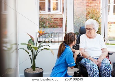Senior Woman Sitting In Motorized Wheelchair Talking With Nurse In Retirement Home
