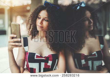 A Charming Curly Caucasian Girl Is Typing A Message On Her Smartphone While Leaning Against Glass Wa