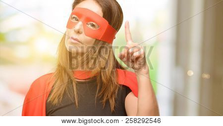 Middle age super hero woman wearing red mask and cape happy and surprised cheering expressing wow gesture pointing up