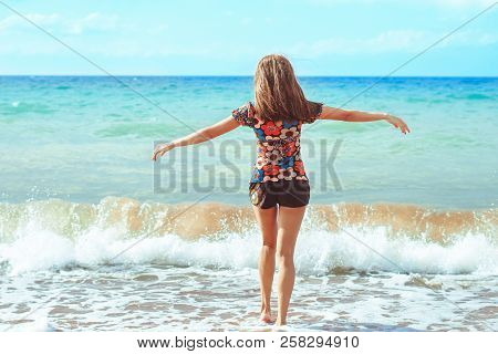 A Young Girl In Shorts Stands With Her Back Against The Background Of Big Waves