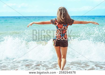 A Young Girl In Shorts Stands With Her Back Against The Background Of Big Waves
