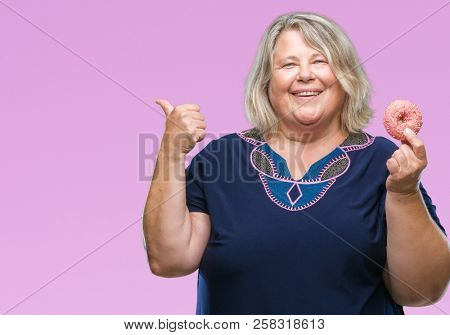Senior plus size caucasian woman eating sugar donut over isolated background pointing and showing with thumb up to the side with happy face smiling