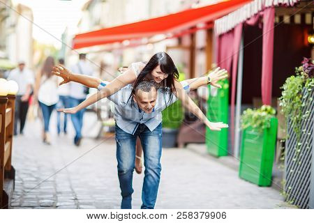 Young Man Piggybacking His Happy Girlfriend With Raised Hand. Cheerful Lovers Dreaming, Walk The Cit