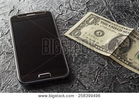 Phone And One Dollar On A Dark, Heterogeneous Background. Black, Brown, Gray, Color.