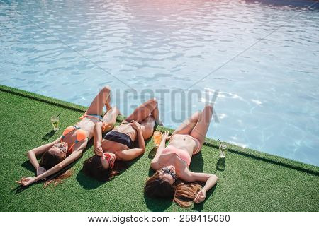 Three Models Are Lying On Grass. They Holds Legs In Water And Get Tan From Sun. Models Stratching An