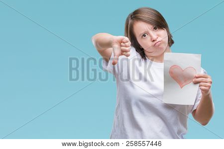 Young adult woman with down syndrome holding red heart card over isolated background with angry face, negative sign showing dislike with thumbs down, rejection concept