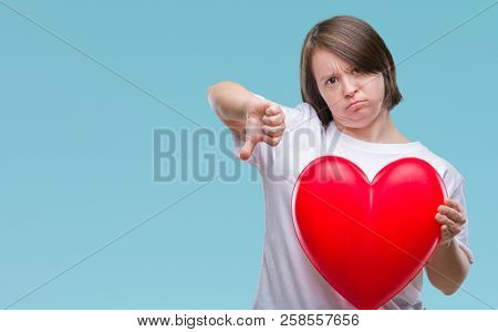 Young adult woman with down syndrome holding red heart over isolated background with angry face, negative sign showing dislike with thumbs down, rejection concept