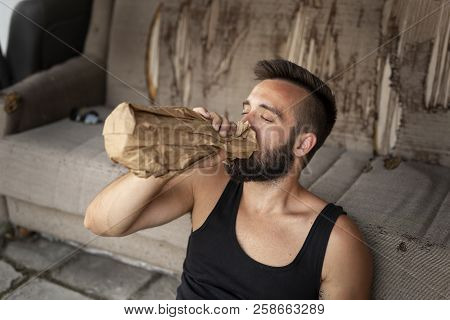 Drunkard Sitting On A Building Rooftop Terrace, Holding A Bottle In A Paper Bag, Drinking