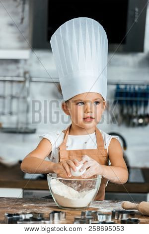 Adorable Child In Aron And Chef Hat Cooking Dough And Looking Away In Kitchen