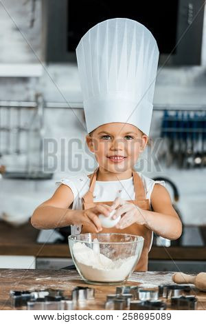Adorable Child In Aron And Chef Hat Cooking Dough And Smiling At Camera