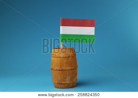 A View On National Flag Of Hungaria On Wooden Stick In Wooden Barrel. Blue Background