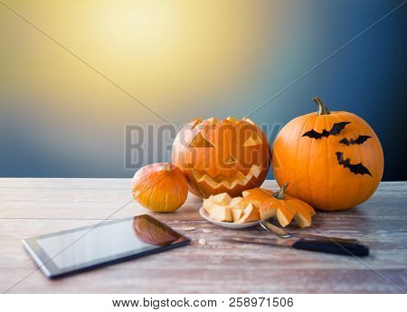 holidays, halloween and technology concept - jack-o-lantern or carved pumpkin with tablet pc computer and knife on wooden table over moonlight background