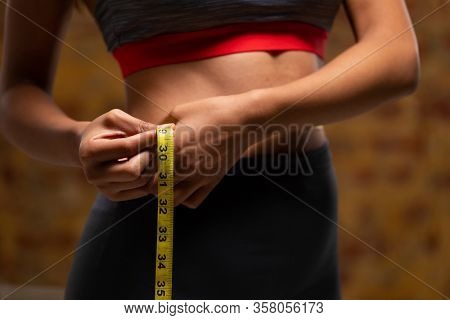 Side view mid section of a fit mixed race woman wearing sportswear and measuring her waist with measuring tape with brick wall in the background.