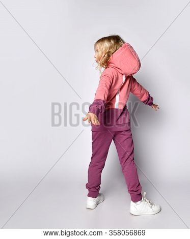 Curly Hair Blonde Kid Girl In Modern Fashion Pink Gray Sportsuit Stands Back To Us With Her Arms Out