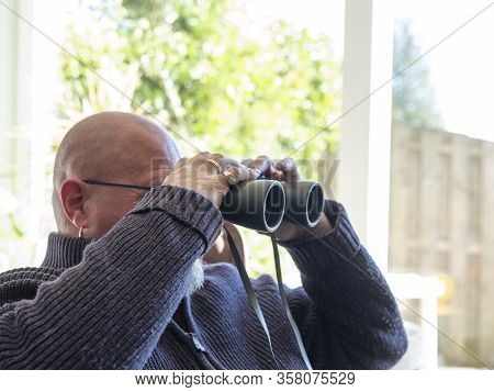 Elderly Man Sittiing, Looking Through A Glass Door With Binoculars As He Watches Something In The Di