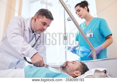 Concentrated mature doctor using stethoscope while examining lungs of patient in hospital room, nurse with ambu mask standing behind bed