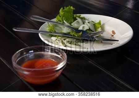 Leftover Lettuce And Onion Rings On White Plate With Fork And Knife Next To A Red Sauce Bar.